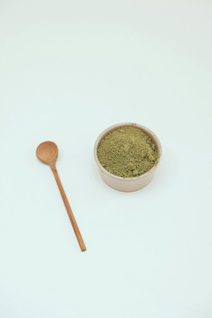 Simple vertical shot of matcha green tea powder in a bowl with a wooden spoon on a white surface.